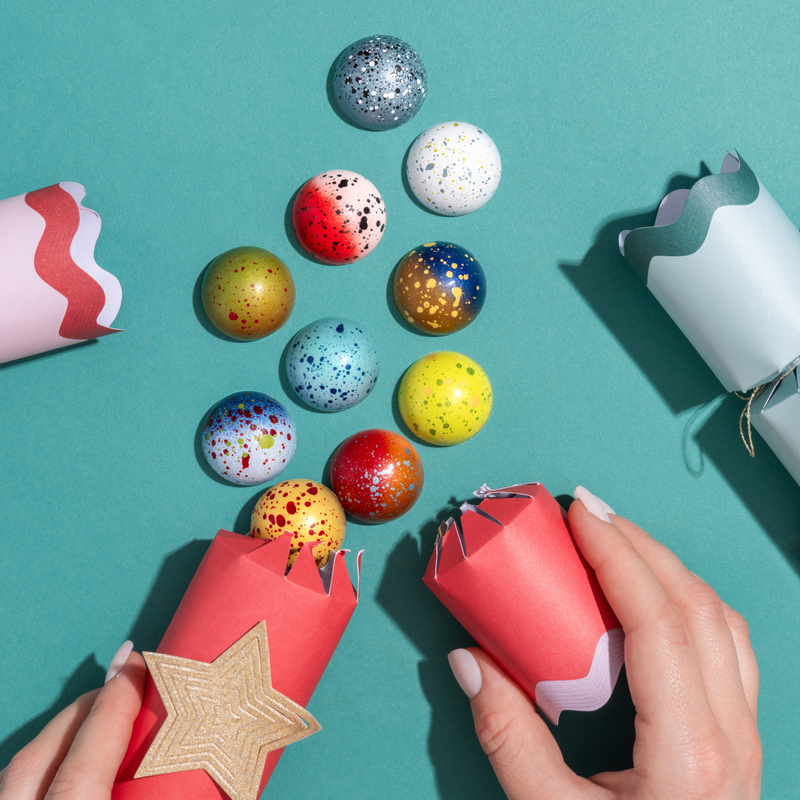 10 chocolates from the holiday collection are spread out on a blue background as if they are the opened contents of a gift. Two hands holding paper gift boxes are shown at the bottom.
